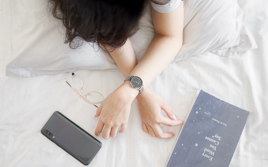 A person sits on a bed with just their arms visible, crossed one over the other. They wear a watch, and one hand reaches to a phone face down on the blanket, the other to the book "Every Word You Cannot Say" by Iain S. Thomas. The colours are light, with the phone, book, person's hair, and an analog watch standing out in dark brown and grey.