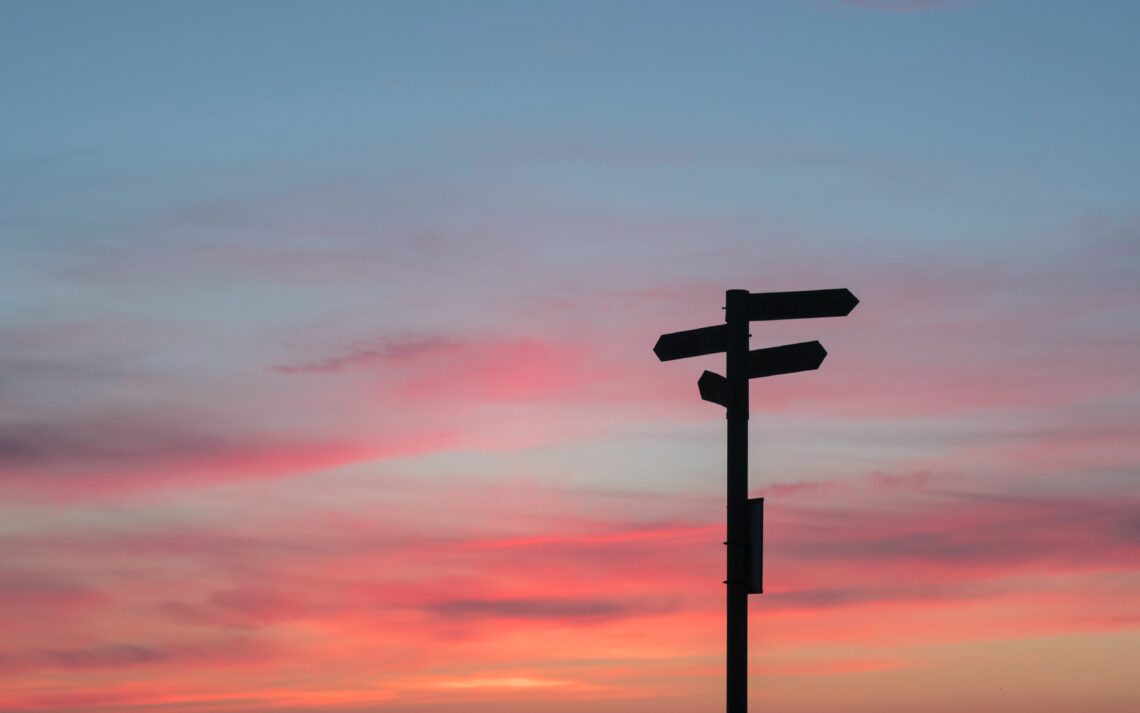 A soft sunset behind the silhouette of a road sign with four directional signs pointing in different directions.